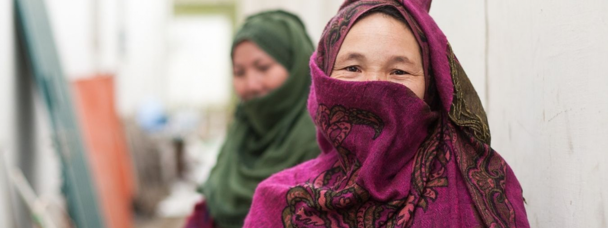 Afghan woman foregrounded wearing a bright pink scarf. Behind her a woman wears a green headscarf,